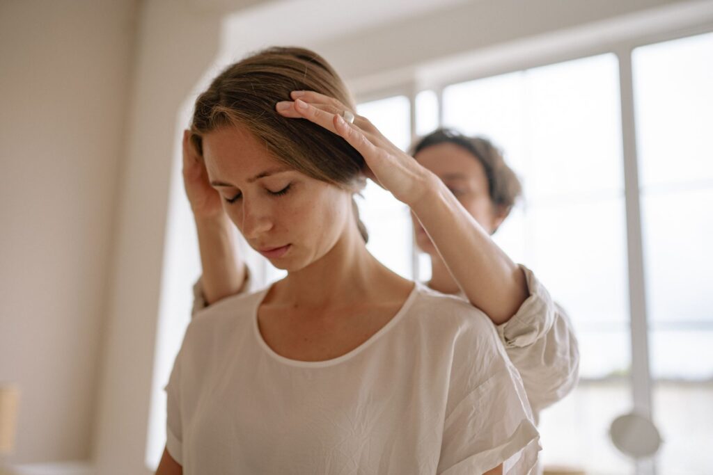 Femme assise, les yeux fermés, pendant que la praticienne pose les mains sur sa tête